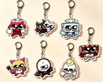 Puede incluir: Una colección de siete llaveros de acrílico con personajes de dibujos animados de ojos grandes. Los diseños incluyen una lámpara roja, dos figuras pequeñas, una caja de pañuelos azul, un gato amarillo, un personaje blanco y un rollo rojo.