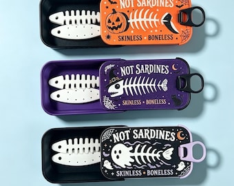 Puede incluir: Tres latas rectangulares con tapa deslizante, cada una con dos formas de esqueleto de pez blancas. La tapa naranja muestra una calabaza, la morada un sombrero de bruja y la negra un fantasma. Todas las tapas muestran el texto NOT SARDINES SKINLESS BONELESS.
