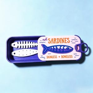 Puede incluir: Un recipiente rectangular morado con tapa deslizante que presenta un gr&aacute;fico de un pez y el texto "not SARDINES" y "SKINLESS + BONELESS". Dentro del recipiente hay dos objetos blancos con forma de pez, uno esqueleto y otro punteado.