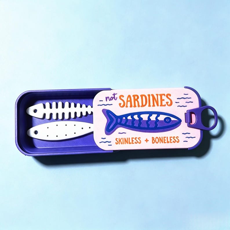 Puede incluir: Un recipiente rectangular morado con tapa deslizante que presenta un gr&aacute;fico de un pez y el texto "not SARDINES" y "SKINLESS + BONELESS". Dentro del recipiente hay dos objetos blancos con forma de pez, uno esqueleto y otro punteado.