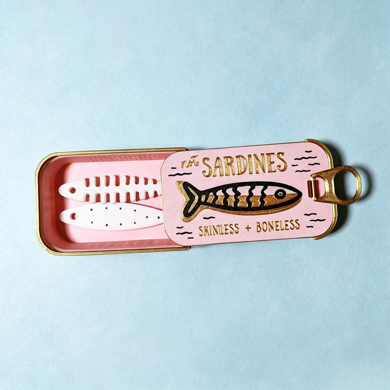 Puede incluir: Una lata de sardinas de metal rosa con borde y anilla dorados. La tapa presenta un gr&aacute;fico de pez en negro y oro con el texto "THE SARDINES" y "SKINLESS + BONELESS". En el interior, se ven dos objetos blancos planos con forma de pez.