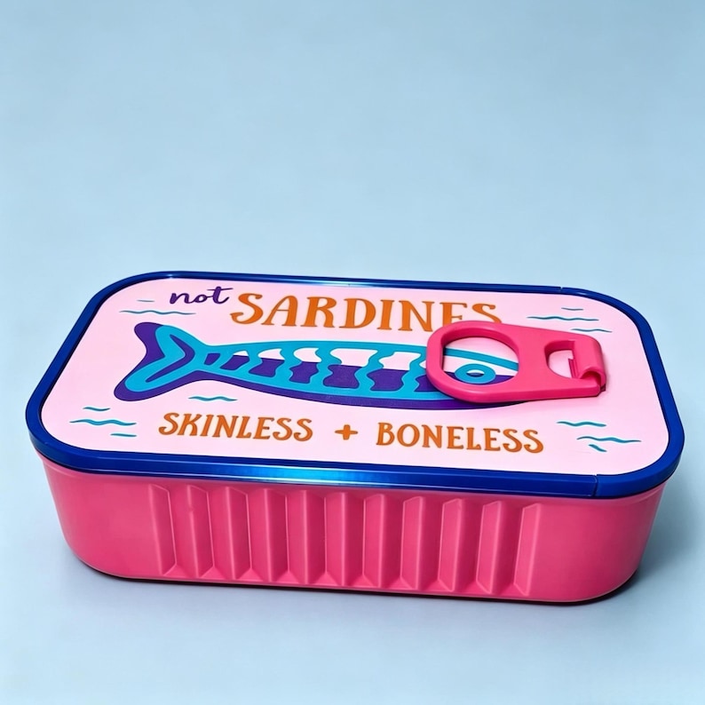 Puede incluir: Un recipiente de almacenamiento rectangular de color rosa con un borde azul oscuro y una tapa con el gr&aacute;fico de un pez azul. El texto de la tapa dice "not SARDINES" y "SKINLESS + BONELESS" en marr&oacute;n, con una anilla rosa en la parte superior.