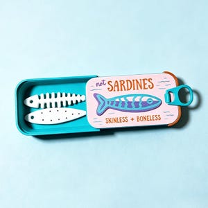 Op de afbeelding: Een talingkleurig rechthoekig blik met een schuifdeksel met een roze achtergrond, een blauwe visillustratie en de tekst "not SARDINES" en "SKINLESS + BONELESS". In het blik liggen twee witte, platte, visvormige objecten met uitsparingen.