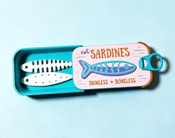 Puede incluir: Una lata rectangular de color cerceta con tapa deslizante que presenta un fondo rosa, una ilustración de un pez azul y el texto "not SARDINES" y "SKINLESS + BONELESS". Dentro de la lata hay dos objetos blancos planos con forma de pez.