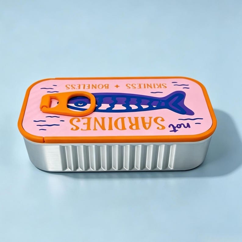 Puede incluir: Un recipiente rectangular de metal con tapa rosa que presenta una anilla naranja, un gr&aacute;fico de pez morado y el texto "not SARDINES" y "SKINLESS + BONELESS". Los lados del recipiente son plateados con estr&iacute;as verticales y un borde naranja.