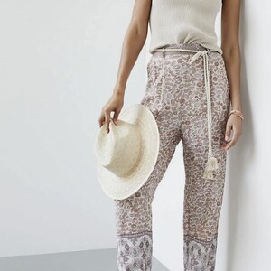 May include: A woman's outfit featuring a cream-colored ribbed tank top, patterned pants with a beige and brown design, and a straw hat. The pants have a decorative border at the hem. The outfit is accessorized with a rope belt and sandals.