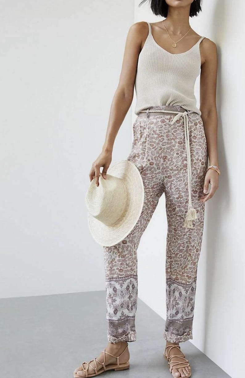 May include: A woman's outfit featuring a cream-colored ribbed tank top, patterned pants with a beige and brown design, and a straw hat. The pants have a decorative border at the hem. The outfit is accessorized with a rope belt and sandals.