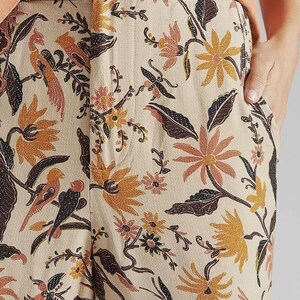 Puede incluir: Pantalones beige con un vibrante estampado floral y de p&aacute;jaros en tonos naranja, marr&oacute;n y negro. Los pantalones tienen cierre de bot&oacute;n y bolsillos. El tema general es ropa casual.