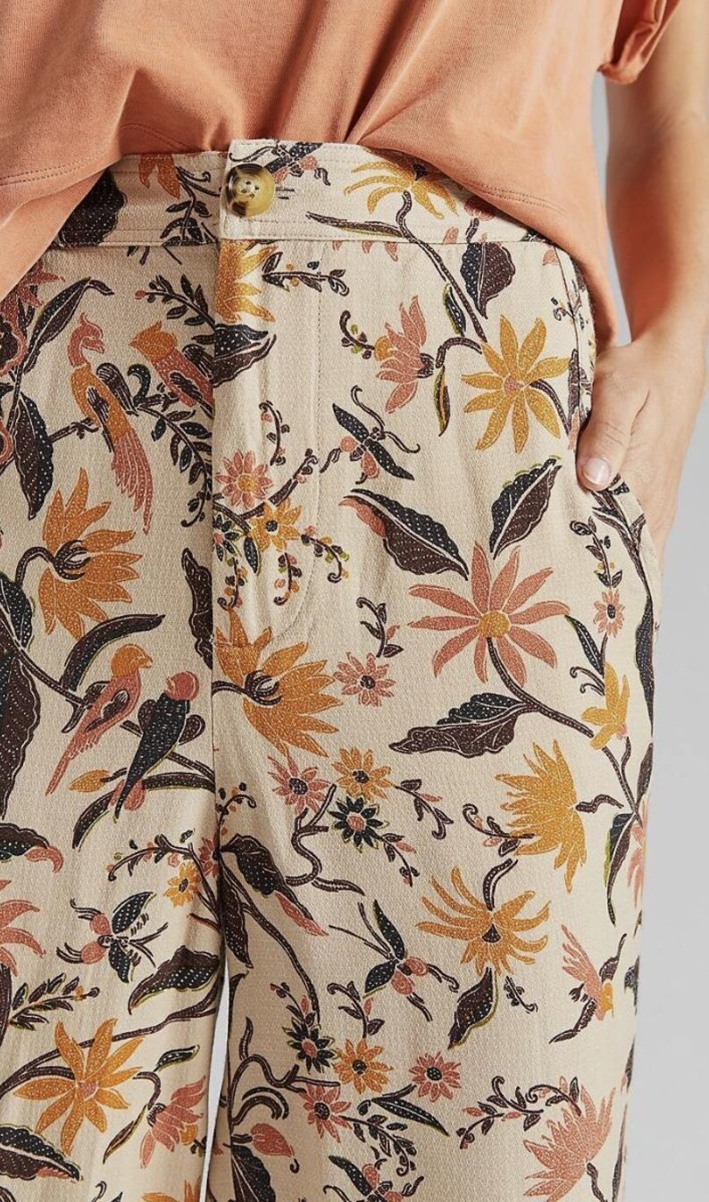 Puede incluir: Pantalones beige con un vibrante estampado floral y de p&aacute;jaros en tonos naranja, marr&oacute;n y negro. Los pantalones tienen cierre de bot&oacute;n y bolsillos. El tema general es ropa casual.