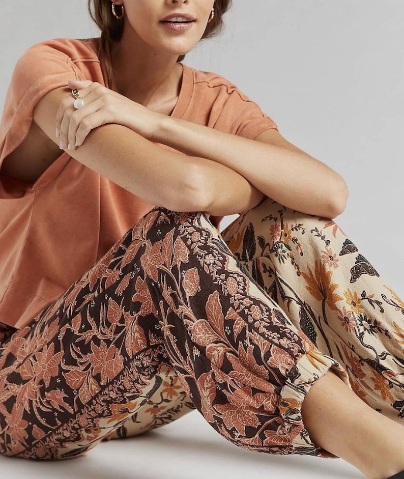 Puede incluir: Una persona con una camiseta color melocot&oacute;n y pantalones con estampado floral. Los pantalones presentan un estampado floral marr&oacute;n oscuro y melocot&oacute;n sobre un fondo crema. Los pantalones tienen pu&ntilde;os el&aacute;sticos en los tobillos.