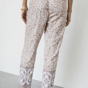 May include: Tan and white patterned pants with a leopard print design. The pants have a decorative border at the ankles with a floral pattern. The pants have a drawstring waist and are worn with sandals.