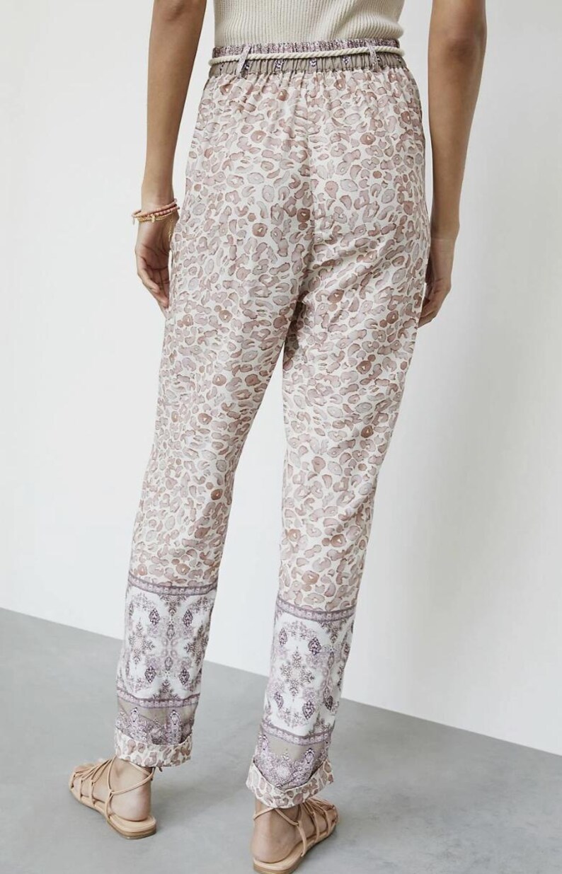 May include: Tan and white patterned pants with a leopard print design. The pants have a decorative border at the ankles with a floral pattern. The pants have a drawstring waist and are worn with sandals.