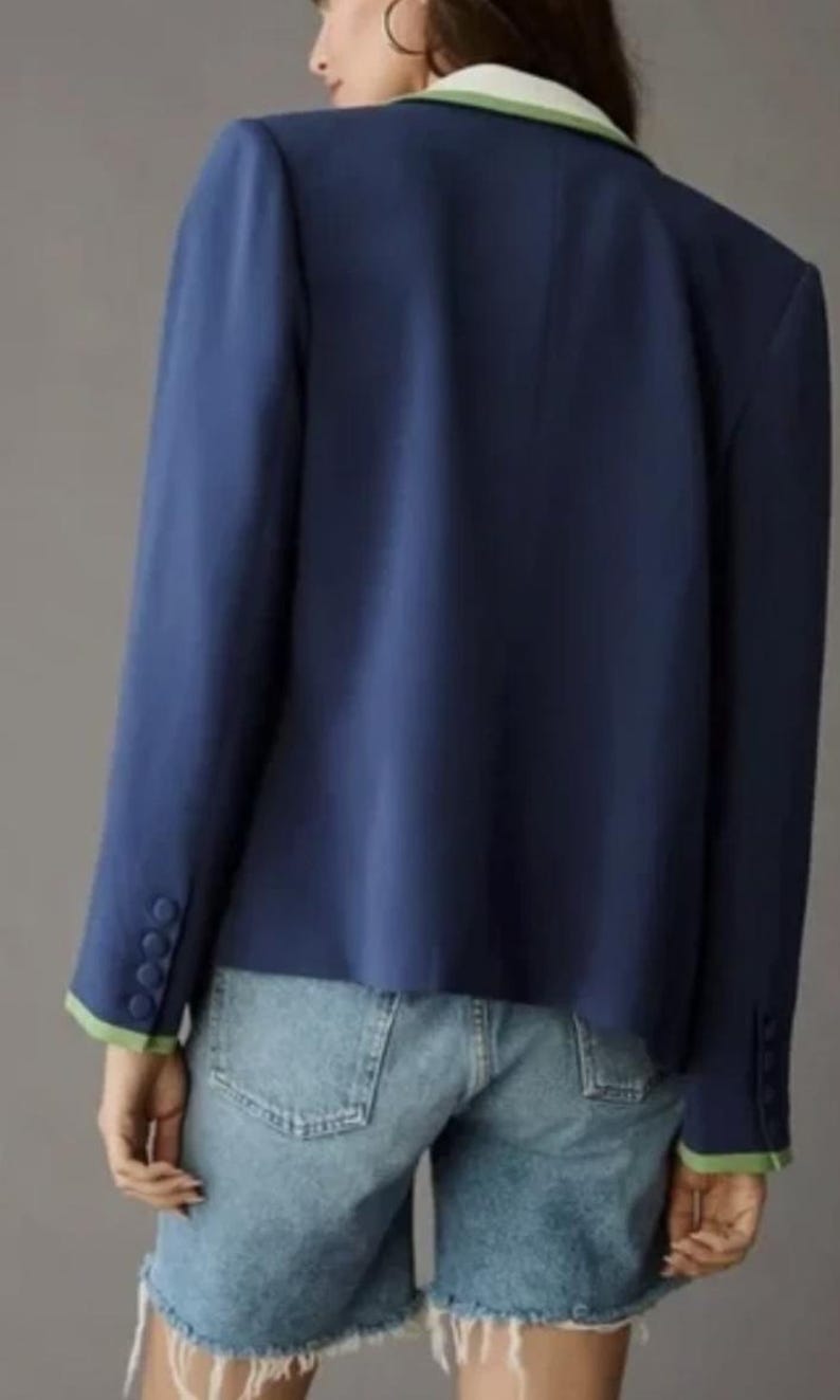 May include: A navy blue blazer with a white and green trim on the collar and cuffs. The blazer has three buttons on each cuff. The model is wearing denim shorts.