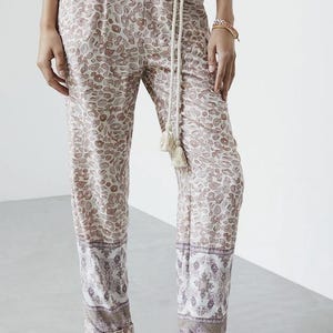 May include: Tan and white patterned pants with a matching belt and rope tie. The pants have a decorative border at the hem and are paired with neutral sandals. The overall style is casual and bohemian.