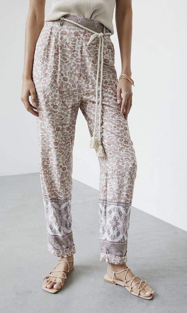May include: Tan and white patterned pants with a matching belt and rope tie. The pants have a decorative border at the hem and are paired with neutral sandals. The overall style is casual and bohemian.