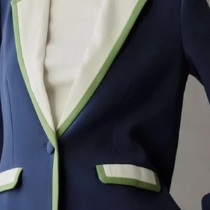 May include: A navy blue blazer with white lapels and pocket trim, accented with a green border. The blazer has a single-button closure and a classic tailored design. The garment is part of a clothing collection.