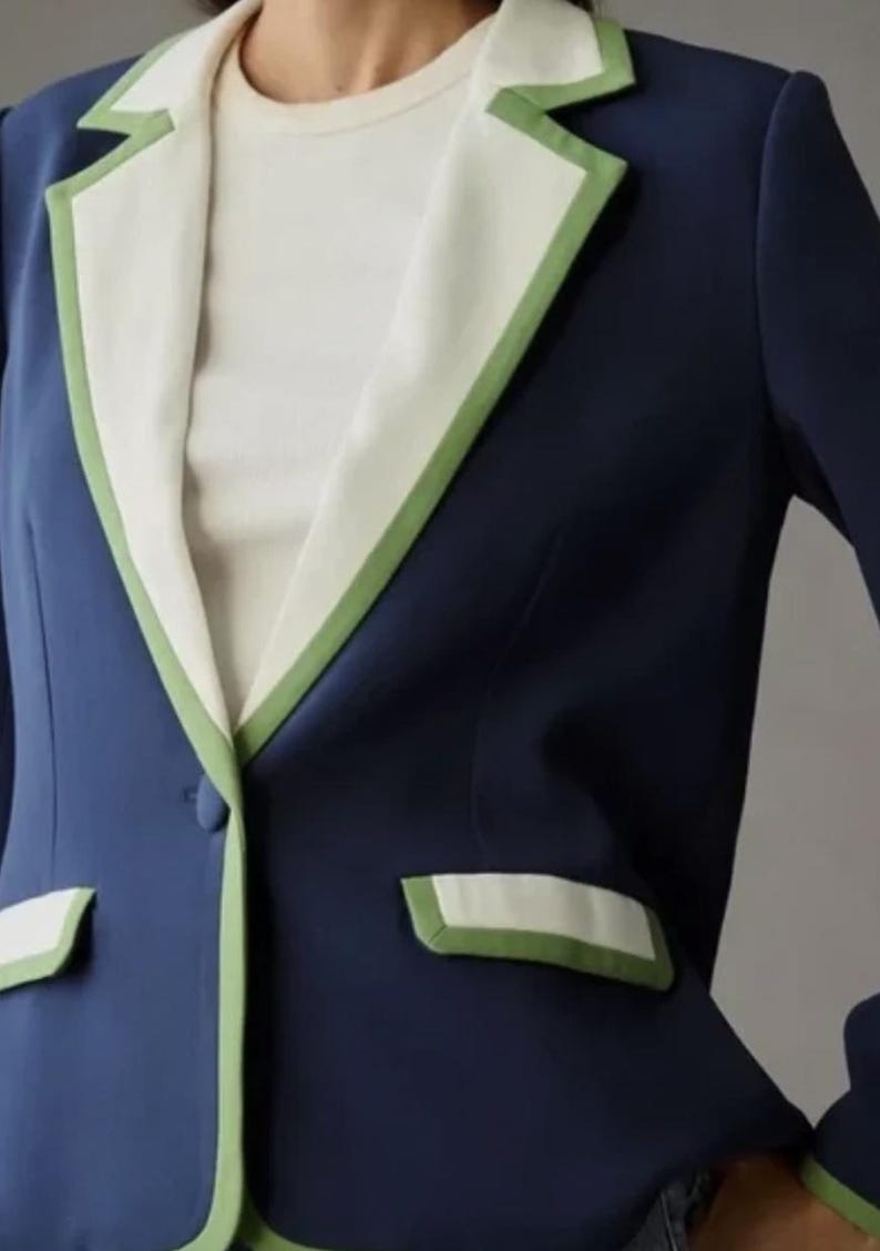 May include: A navy blue blazer with white lapels and pocket trim, accented with a green border. The blazer has a single-button closure and a classic tailored design. The garment is part of a clothing collection.