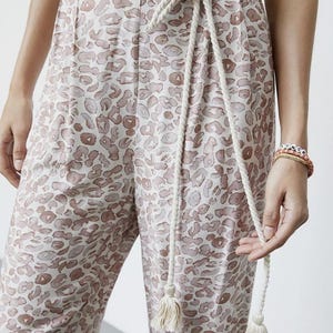 May include: Cream-colored pants with a brown leopard print. The pants have a matching rope belt with tassels. The model is wearing a cream-colored top and bracelets. The pants are made of a soft, lightweight material.