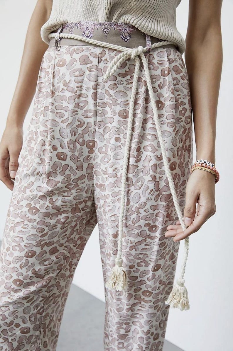 May include: Cream-colored pants with a brown leopard print. The pants have a matching rope belt with tassels. The model is wearing a cream-colored top and bracelets. The pants are made of a soft, lightweight material.