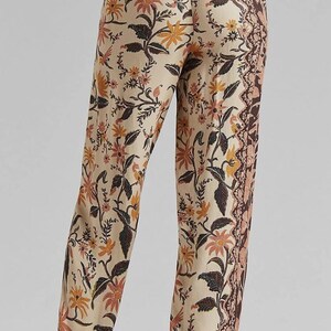Puede incluir: Pantalones beige con estampado floral y borde marr&oacute;n. Los pantalones presentan un patr&oacute;n repetido de flores y hojas en tonos marr&oacute;n, naranja y rosa. Los pantalones est&aacute;n hechos de tela ligera.
