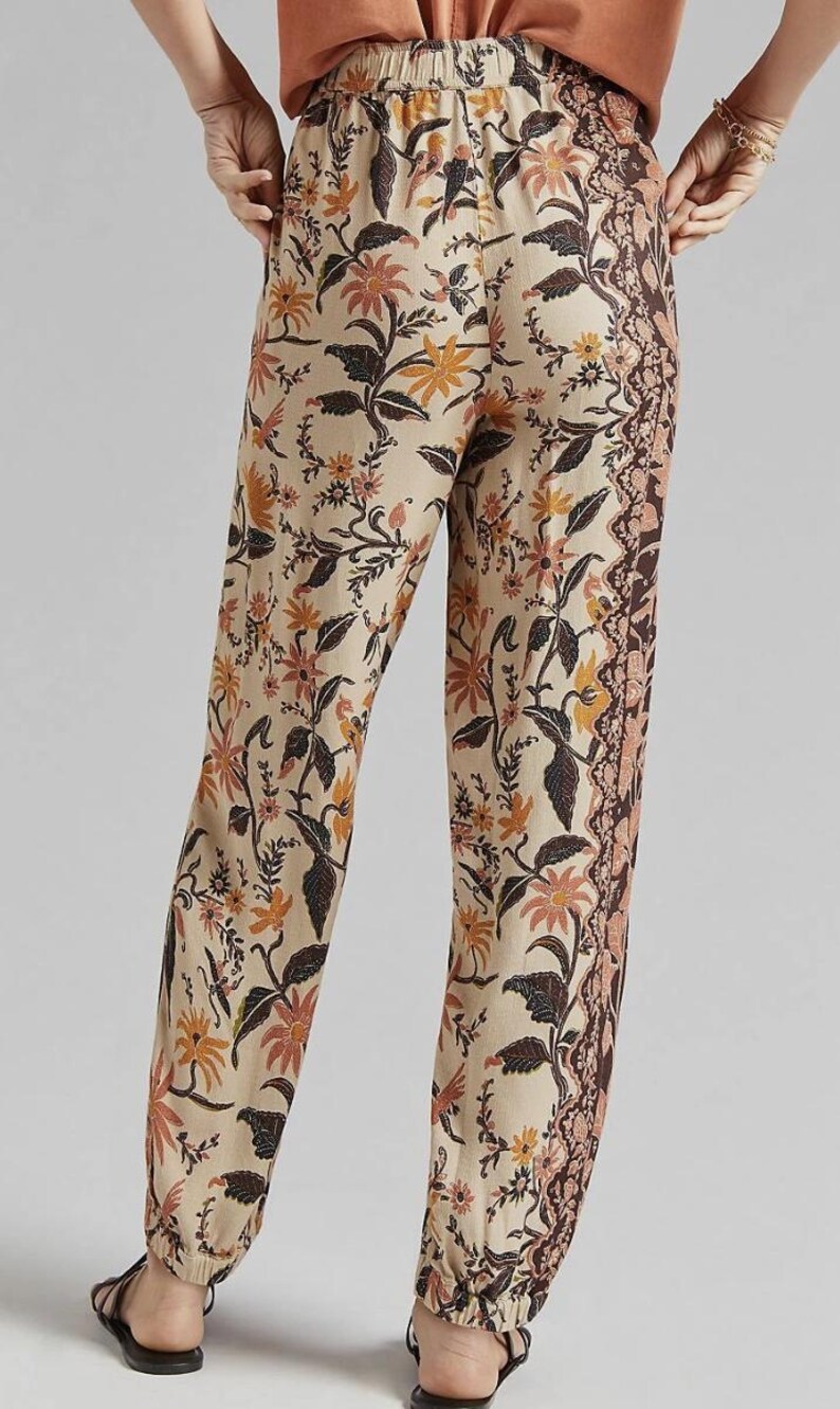 Puede incluir: Pantalones beige con estampado floral y borde marr&oacute;n. Los pantalones presentan un patr&oacute;n repetido de flores y hojas en tonos marr&oacute;n, naranja y rosa. Los pantalones est&aacute;n hechos de tela ligera.