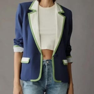 May include: A navy blue blazer with white lapels and green trim. The blazer has a single button closure and pocket details. The sleeves are cuffed with a striped pattern. The model is wearing a white top and blue jeans.