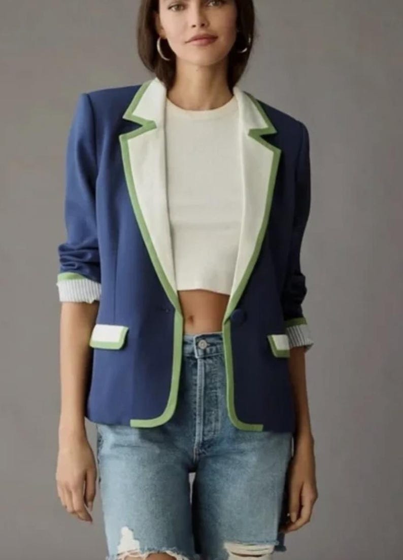 May include: A navy blue blazer with white lapels and green trim. The blazer has a single button closure and pocket details. The sleeves are cuffed with a striped pattern. The model is wearing a white top and blue jeans.