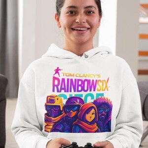 May include: White hoodie featuring the text "Tom Clancy's Rainbow Six Siege" in colorful lettering, with a graphic of four characters. The hoodie is worn by a person holding a black video game controller.