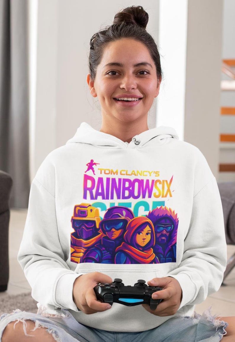 May include: White hoodie featuring the text "Tom Clancy's Rainbow Six Siege" in colorful lettering, with a graphic of four characters. The hoodie is worn by a person holding a black video game controller.