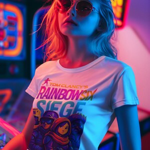 May include: A white crop top featuring the text "TOM CLANCY'S RAINBOW SIX SIEGE" with a graphic of three characters. The image is set in a neon-lit arcade environment, with the subject wearing sunglasses.