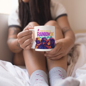May include: White ceramic mug featuring the text "Tom Clancy's Rainbow Six Siege" with a graphic of four characters. The mug has a white handle and is held by a person wearing gray socks.