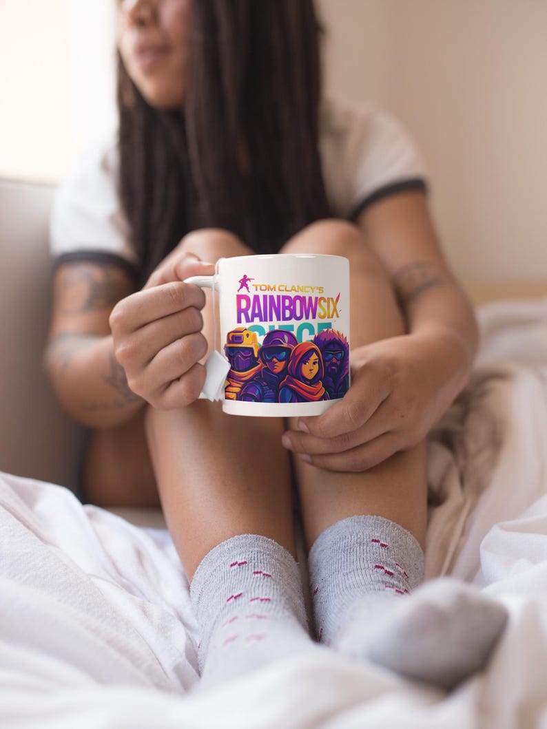 May include: White ceramic mug featuring the text "Tom Clancy's Rainbow Six Siege" with a graphic of four characters. The mug has a white handle and is held by a person wearing gray socks.