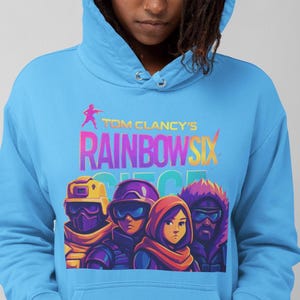 May include: Light blue hoodie featuring the text "Tom Clancy's Rainbow Six" above a graphic of four characters. The graphic is in vibrant colors, and the hoodie has a drawstring hood and a front pocket.