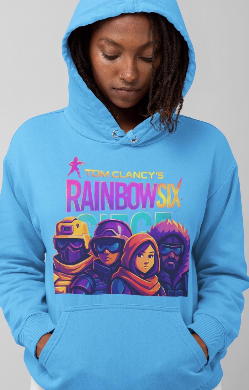 May include: Light blue hoodie featuring the text "Tom Clancy's Rainbow Six" above a graphic of four characters. The graphic is in vibrant colors, and the hoodie has a drawstring hood and a front pocket.