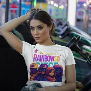May include: White t-shirt featuring the text "Tom Clancy's Rainbow Six Siege" in a rainbow gradient. The graphic includes four stylized characters in tactical gear. The shirt is being worn.