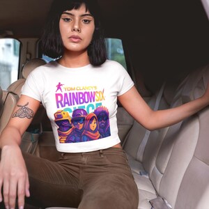 May include: White crop top featuring the text "Tom Clancy's Rainbow Six Siege" in a vibrant, colorful design. The graphic includes stylized characters in tactical gear. The shirt is worn by a person in a car.