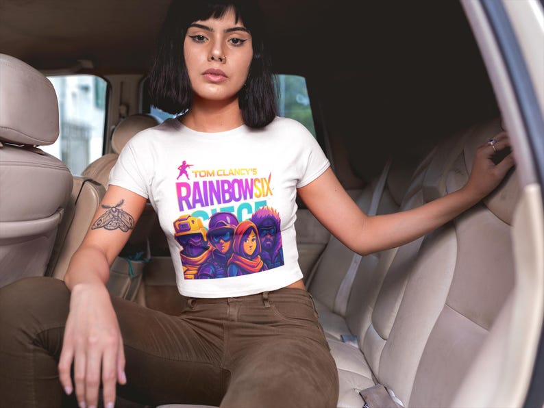 May include: White crop top featuring the text "Tom Clancy's Rainbow Six Siege" in a vibrant, colorful design. The graphic includes stylized characters in tactical gear. The shirt is worn by a person in a car.