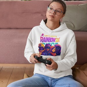 May include: White hoodie featuring the text "Tom Clancy's Rainbow Six Siege" in a colorful design. The hoodie has a graphic of characters from the game. The person in the image is wearing blue jeans and holding a black game controller.