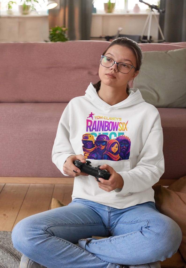 May include: White hoodie featuring the text "Tom Clancy's Rainbow Six Siege" in a colorful design. The hoodie has a graphic of characters from the game. The person in the image is wearing blue jeans and holding a black game controller.