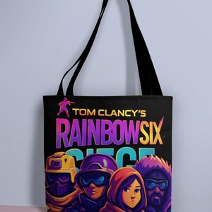 May include: Black tote bag featuring the text "TOM CLANCY'S RAINBOW SIX SIEGE" in vibrant colors. The bag displays a graphic of several characters in tactical gear. The bag has black handles.