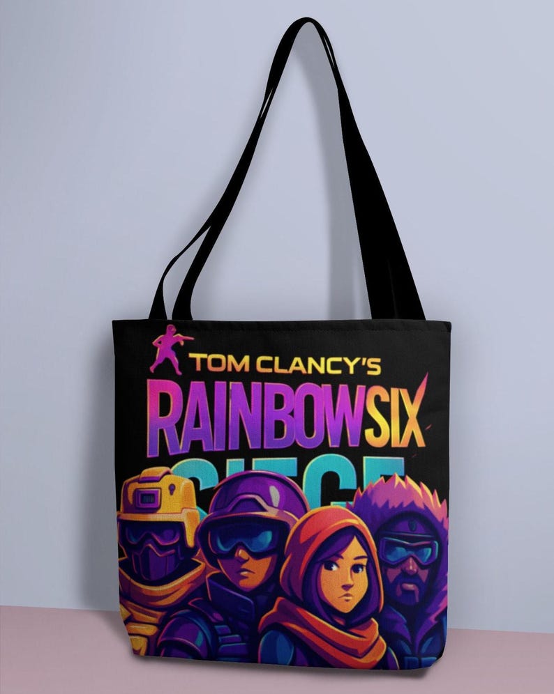 May include: Black tote bag featuring the text "TOM CLANCY'S RAINBOW SIX SIEGE" in vibrant colors. The bag displays a graphic of several characters in tactical gear. The bag has black handles.