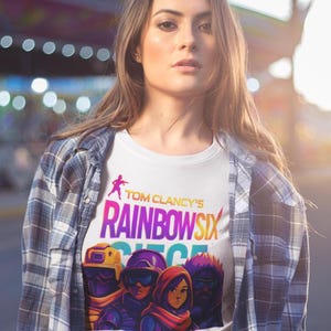 May include: White t-shirt featuring the text "Tom Clancy's Rainbow Six Siege" in a colorful design. The shirt also displays a graphic of four characters. The person is wearing a plaid shirt.
