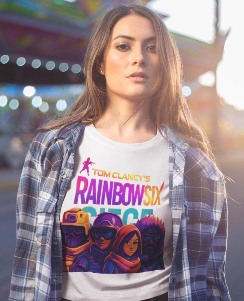 May include: White t-shirt featuring the text "Tom Clancy's Rainbow Six Siege" in a colorful design. The shirt also displays a graphic of four characters. The person is wearing a plaid shirt.