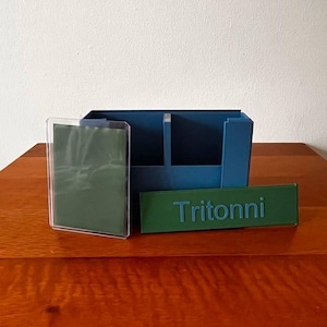May include: A teal desk organizer with two compartments, a clear acrylic display, and a green nameplate that reads "Tritonni". The items are arranged on a wooden surface against a white wall.