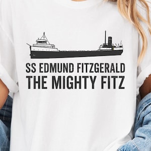 Peut inclure: T-shirt blanc avec un dessin noir d'un navire et le texte "SS Edmund Fitzgerald The Mighty Fitz". Le t-shirt a des manches courtes et un col rond.