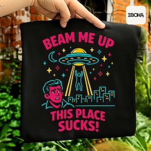 Può includere: Borsa tote nera con una grafica umoristica di un rapimento alieno. Il design include il testo "BEAM ME UP THIS PLACE SUCKS!" con una persona che viene rapita, un paesaggio urbano, stelle e una luna.