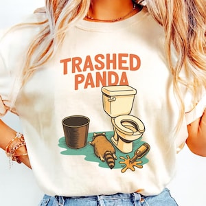 May include: A cream-coloured t-shirt with the text "Trashed Panda" in orange. The graphic shows a raccoon, a toilet, a bin, and a spilled bottle. The raccoon is lying on the floor next to the toilet.