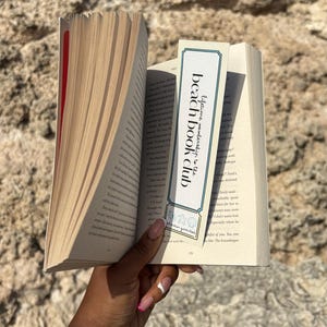 Könnte beinhalten: Ein offenes Buch mit einem Lesezeichen, auf dem "beach book club" steht. Das Lesezeichen ist weiß und blau mit einem Ticket-Design und kleinen Illustrationen. Die Seiten des Buches sind cremefarben, und der Einband ist rot.