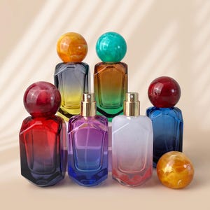Op de afbeelding: Een collectie van zes glazen parfumflesjes met geometrische lichamen en grote bolvormige doppen. De flesjes hebben levendige kleurverlopen in rood, blauw, paars en amber, met goudkleurige verstuivers op de openstaande flesjes.