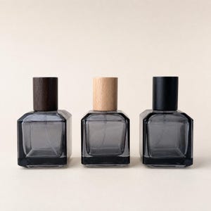 Può includere: Tre flaconi di profumo in vetro grigio con basi quadrate e tappi in legno o neri. I tappi sono marrone scuro, legno chiaro e neri. I flaconi sono disposti in fila su uno sfondo neutro.